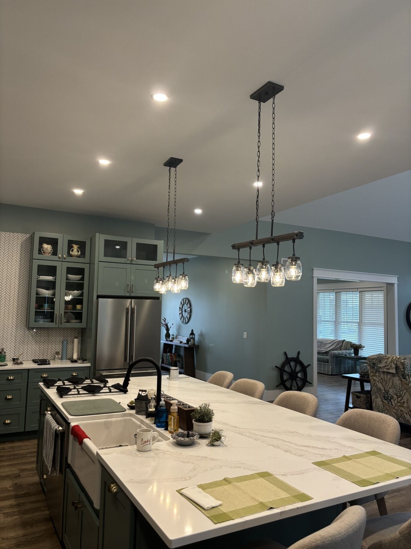 Modern kitchen with green cabinets, marble island, two pendant lights, stainless steel appliances, and a view into the living room. Items are neatly arranged on the countertop.