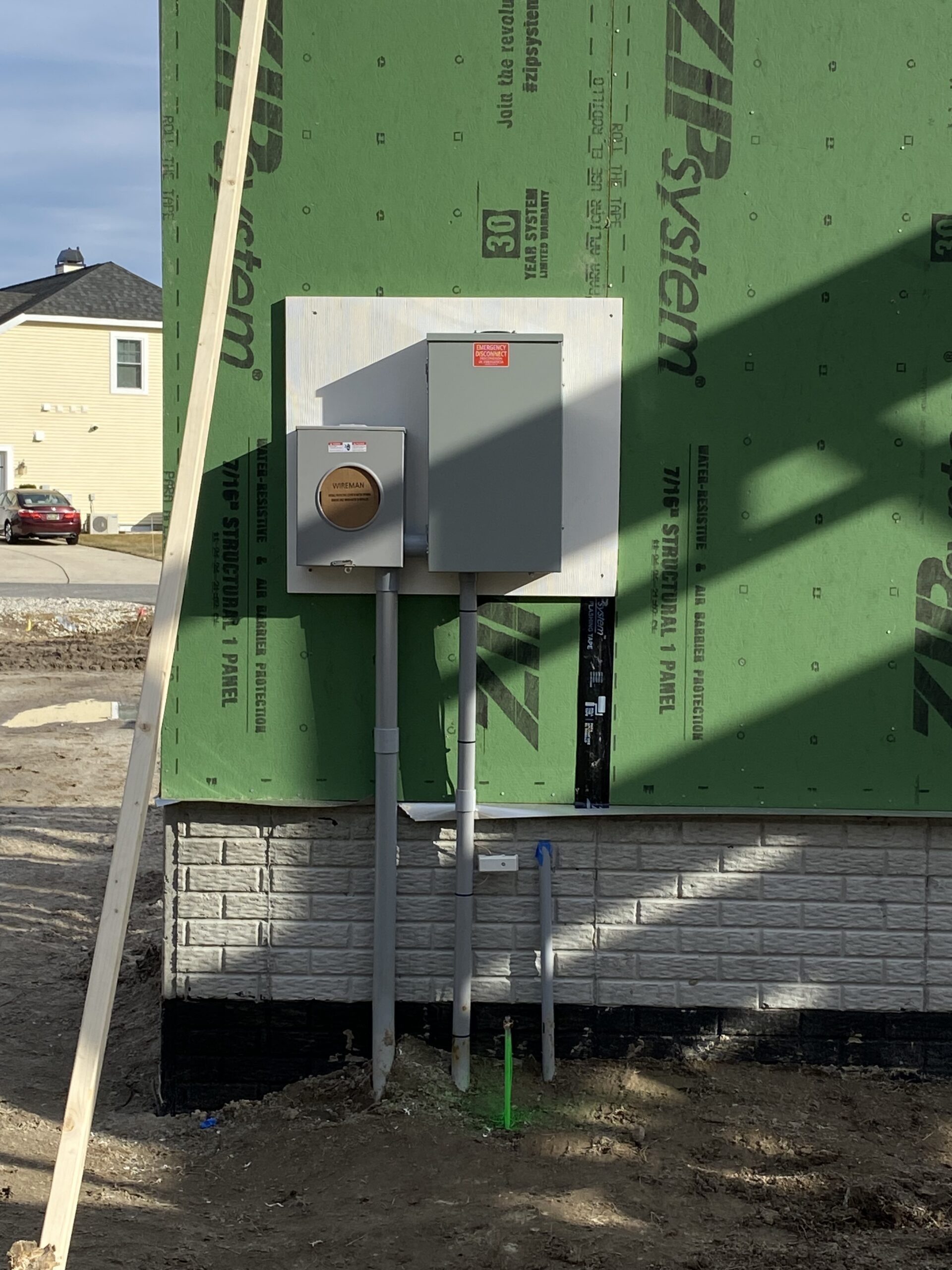Electrical meter and breaker box installed on the exterior green sheathing of a house under construction, with conduit pipes extending to the ground.