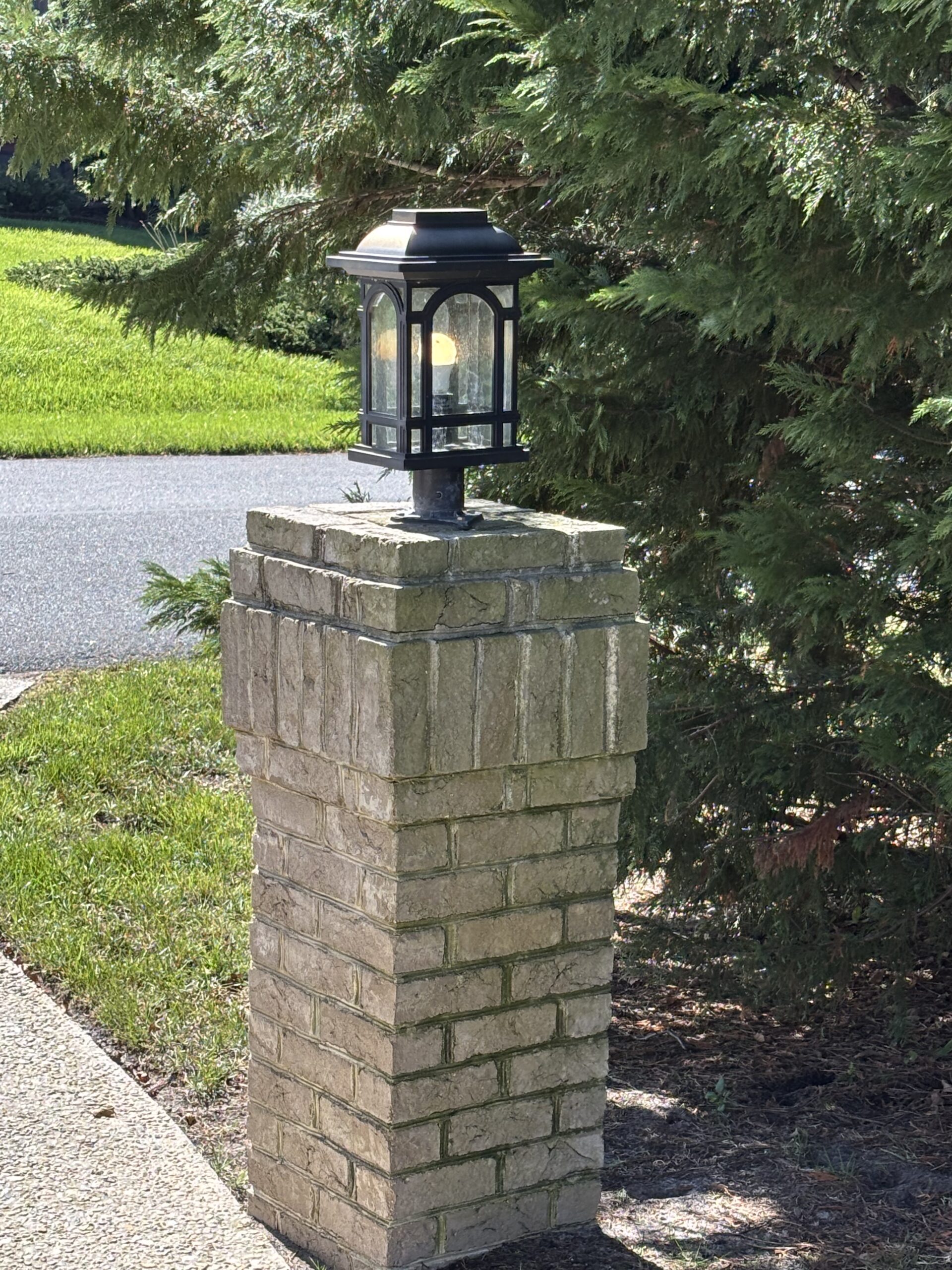 A black metal lantern sits atop a square brick pillar next to a sidewalk, with green grass and trees in the background.
