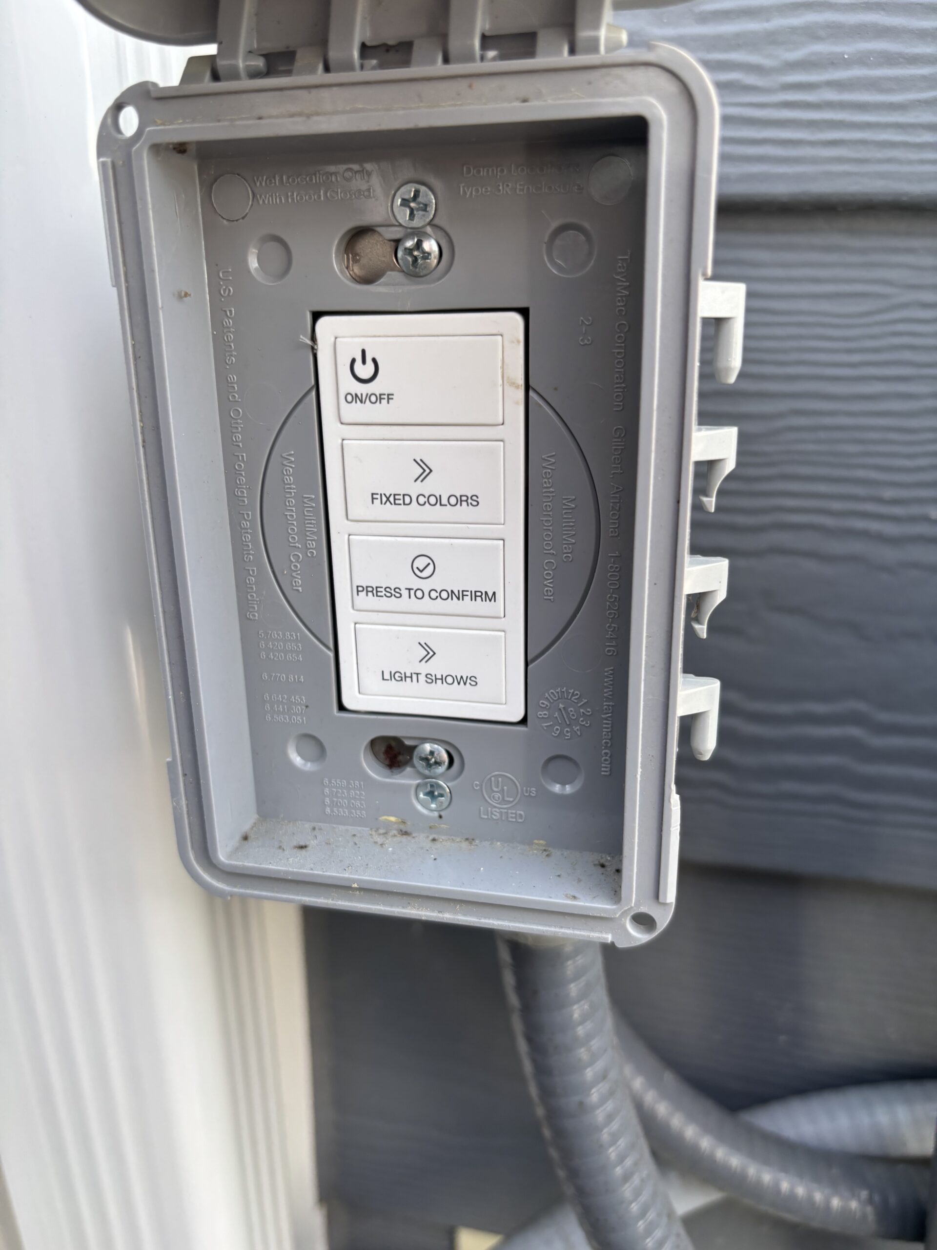 Outdoor electrical control box with buttons labeled ON/OFF, FIXED COLORS, PRESS TO CONFIRM, and LIGHT SHOWS, mounted on a wall with cables below.